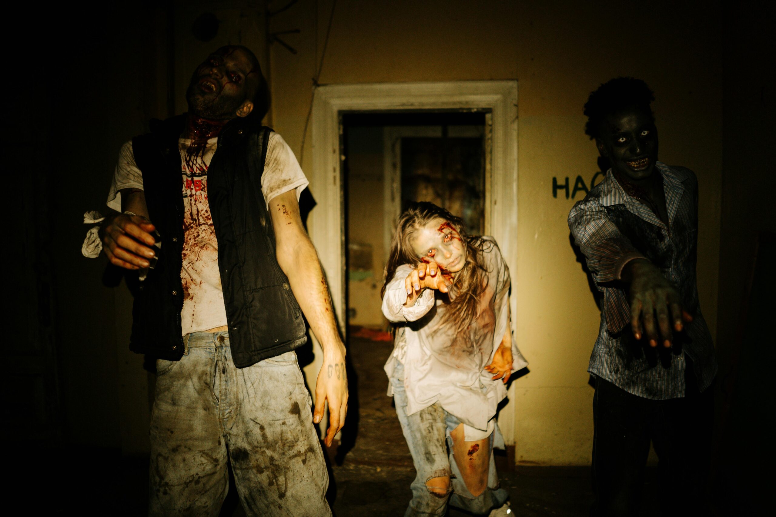 Three costumed zombies outstretch their hands in a dimly lit, abandoned house setting.